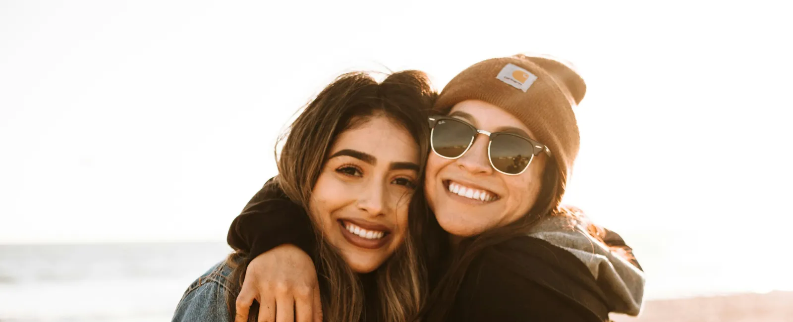Two women smiling and hugging on a sunny beach, one wearing sunglasses and a beanie, the other a denim jacket.