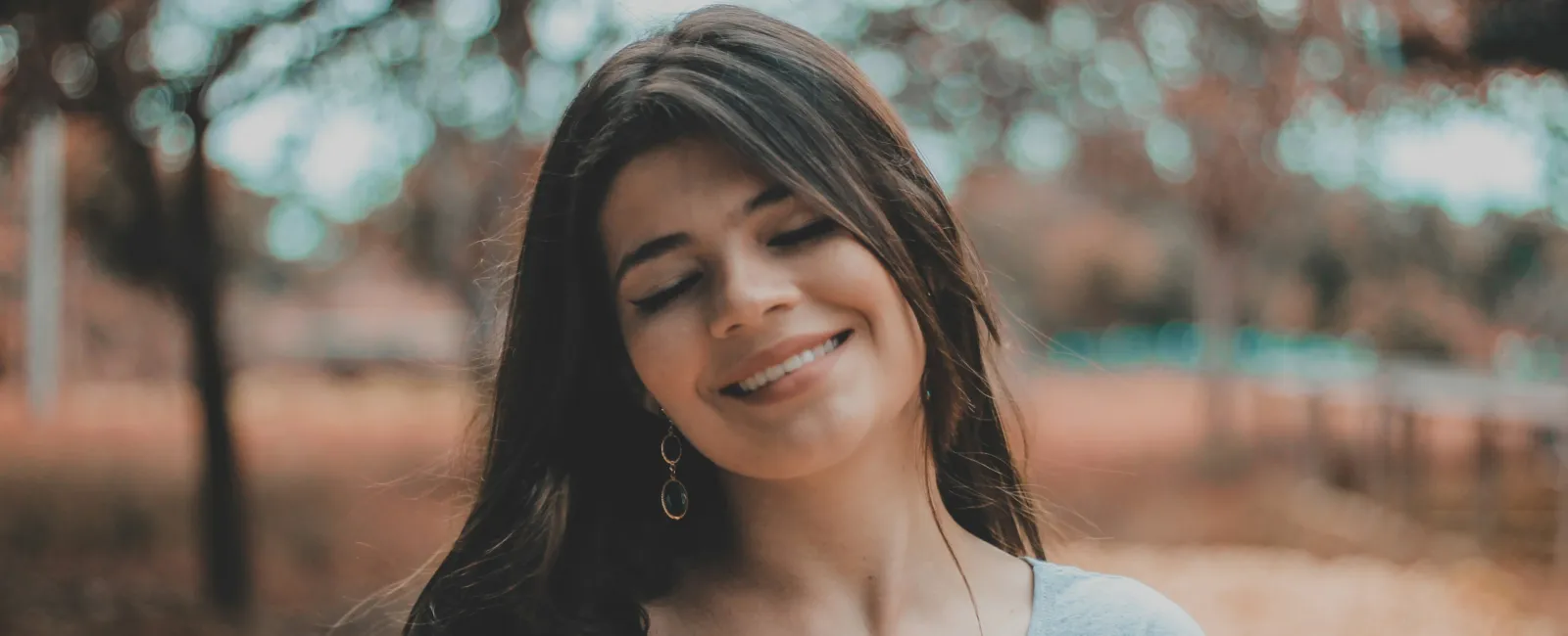 Smiling woman with long dark hair wearing a light blue shirt stands outdoors in a blurred natural setting.
