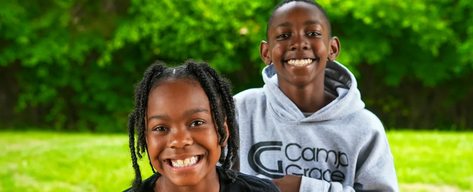 Two smiling children outdoors with vibrant greenery, one in a Camp Grace hoodie and the other in a black shirt.