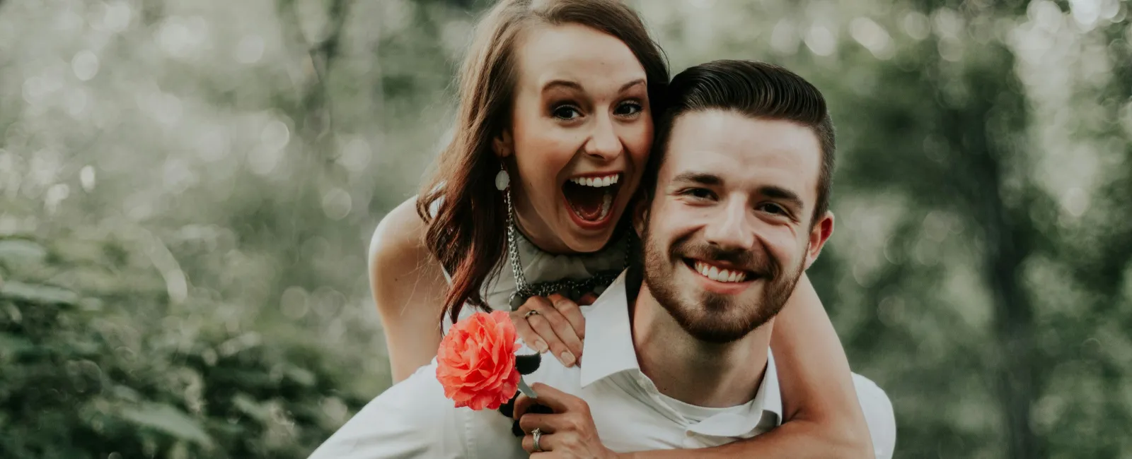 Happy couple outdoors with woman playfully hugging man from behind holding a red flower surrounded by greenery