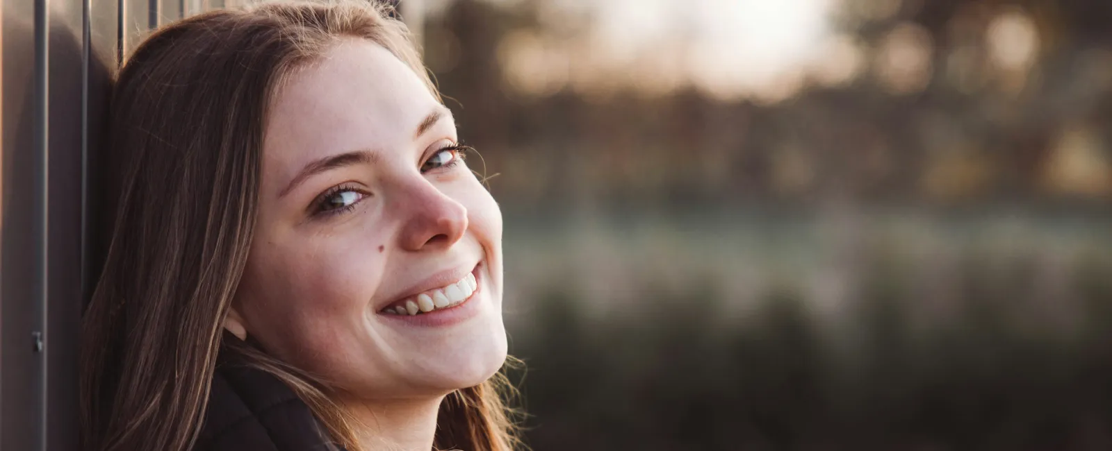 Young woman with long hair smiling outdoors leaning against a wall during golden hour.
