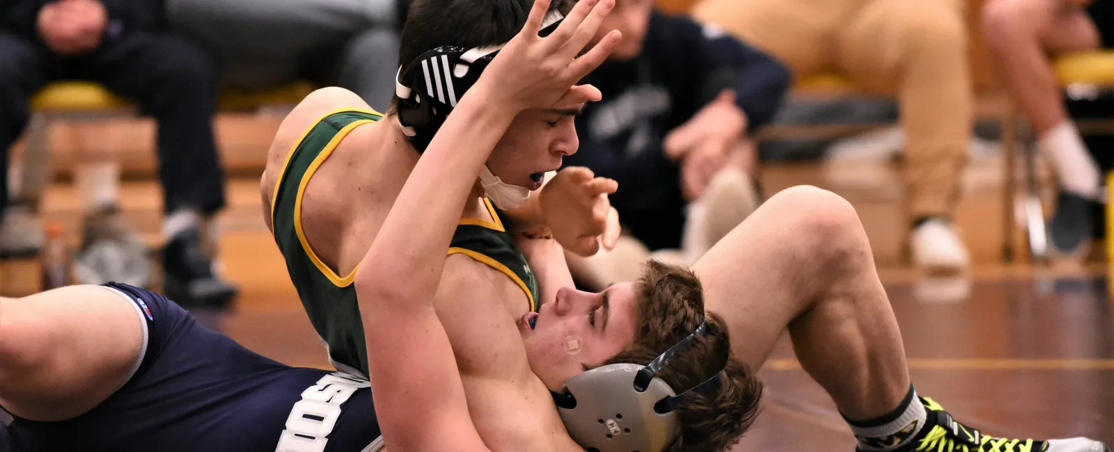 Two high school wrestlers in protective headgear compete closely on a gym wrestling mat during a match.