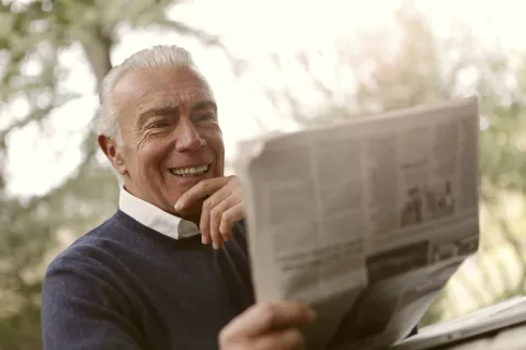 Smiling elderly man reading a newspaper outdoors with natural light and greenery in the background.