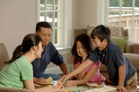 Family of four smiling and playing a board game together around a wooden table in a bright living room