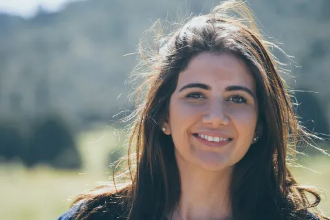 Smiling woman with long dark hair outdoors in natural sunlight with blurred green background.