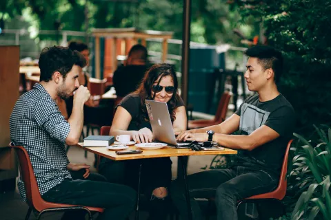 Three people collaborating outdoors at a cafe table with a laptop, coffee, and books in a green garden setting.