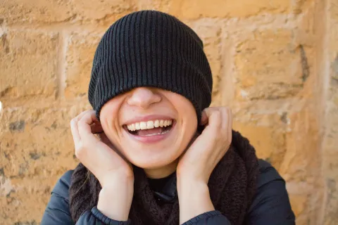 Smiling person with black beanie pulled over eyes laughing against a beige stone wall.