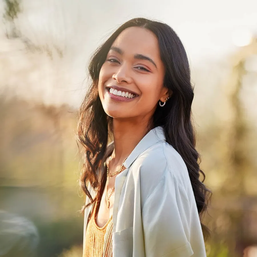 Smiling young woman with long dark hair wearing a light jacket and gold necklace in soft sunlight outdoors.