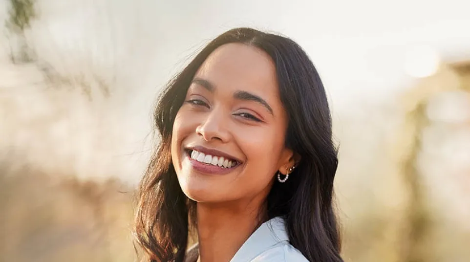 Smiling young woman with long dark hair wearing a light jacket and gold necklace in soft sunlight outdoors.