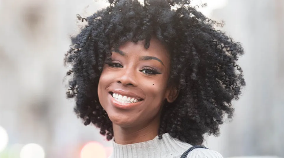 Smiling woman with natural curly hair wearing a light ribbed sweater and carrying a black shoulder bag outdoors.