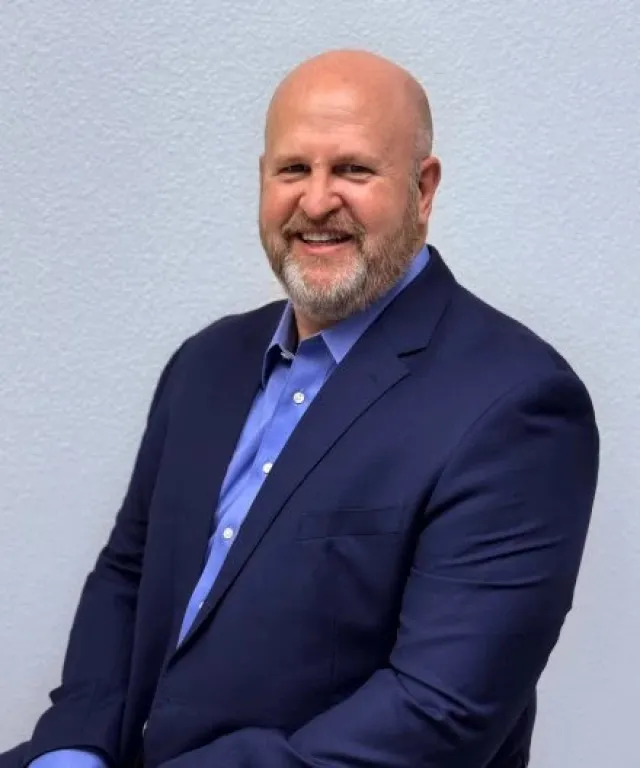 Smiling bald man with beard wearing a navy suit and blue shirt against a plain light background